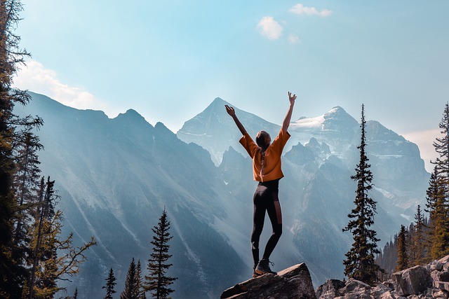 Femme de dos debout sur un rocher qui lève les bras, en fond une montagne enneigée et des arbres autour d'elle