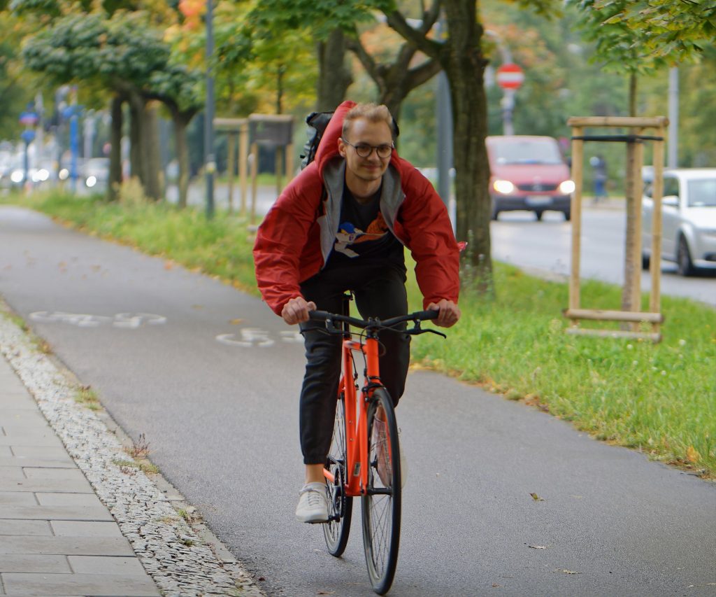 homme blond à vélo en ville, avec une veste rouge et des lunettes